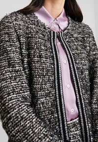 Svartvit texturerad cardigan med en plysch, vävd yta och en svart och silverkant, bärs över en ljuslila satinblus.