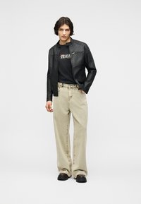 Young man wearing black leather jacket, black Nirvana t-shirt, beige wide-leg jeans, and black shoes, standing against a plain background.