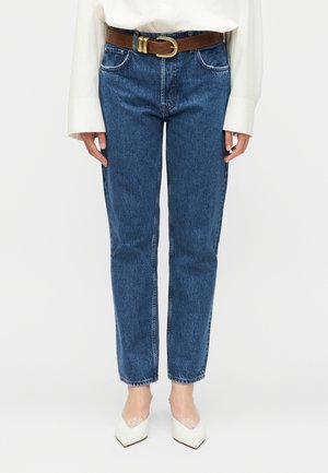 Kvinde iført højtaljede blå jeans med brunt bælte, hvid bluse med lange ærmer og hvide spidse hæle med blonder.