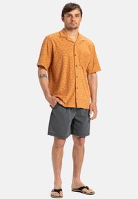 Short-sleeve orange shirt with a textured pattern, black buttons, and a chest pocket, paired with gray shorts and black flip-flops.