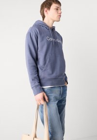 Felpa con cappuccio in blu tenue con tasca frontale, coulisse e logo bianco; abbinata a jeans blu chiaro e una borsa tote beige.