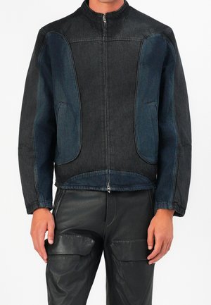 Hombre que lleva una chaqueta negra con cremallera y paneles azul oscuro en las mangas y el frente, combinada con pantalones de cuero negros con bolsillos grandes.