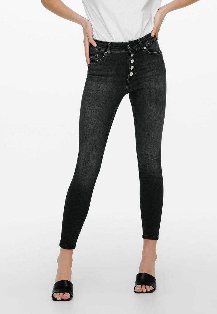 Femme portant un jean skinny noir taille haute avec braguette à boutons, un haut blanc et des sandales noires ouvertes aux orteils, debout les mains sur les hanches.