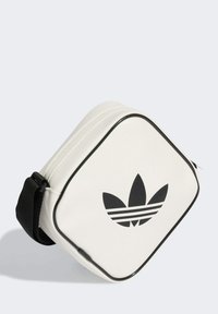 Bolsa rectangular blanca con un acabado texturizado, cremallera negra y un prominente logo de trébol negro de Adidas en la parte frontal. Correa negra adjunta.