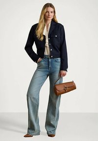 Mujer vestida con una chaqueta azul marino, blusa blanca, jeans de pierna ancha azules, zapatos marrones y sosteniendo un bolso de cuero marrón por su asa.