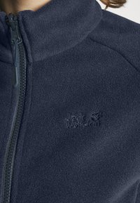 Giacca in pile blu navy con colletto alto, zip completa e logo ricamato. Tessuto liscio con un motivo di tessuto uniforme. Design leggero.