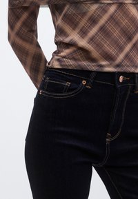 Vaqueros azul oscuro con costuras naranjas, con un diseño clásico de cinco bolsillos y cintura de altura media, combinados con una camisa a cuadros marrón.