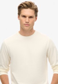 Witte lange mouwen crewneck t-shirt gemaakt van zachte stof, gepresenteerd op een neutrale achtergrond, met opgerolde mouwen en een eenvoudig ontwerp.