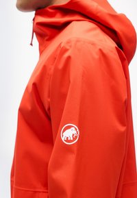 Person iført en knaldorange hættejakke med en hvid mammutlogo på overarmen mod en ensartet baggrund.