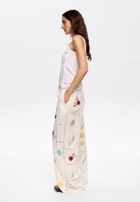 Cremfarbene Baumwoll-Hose mit weitem Bein, verziert mit mehrfarbiger Blumenstickerei und Spitzenakzenten, kombiniert mit einem eng anliegenden weißen Tanktop.