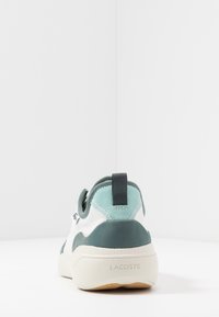 Baskets Lacoste présentant un design blanc et turquoise, des textures en cuir lisse et en daim, une forme arrondie et un marquage discret sur la semelle.