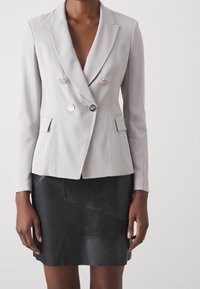 Blazer grigio doppiopetto con bottoni argentati, rever a punta e tasche frontali, abbinato a una gonna di pelle nera con motivo geometrico.