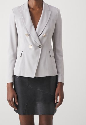 Blazer grigio doppiopetto con bottoni argentati, rever a punta e tasche frontali, abbinato a una gonna di pelle nera con motivo geometrico.
