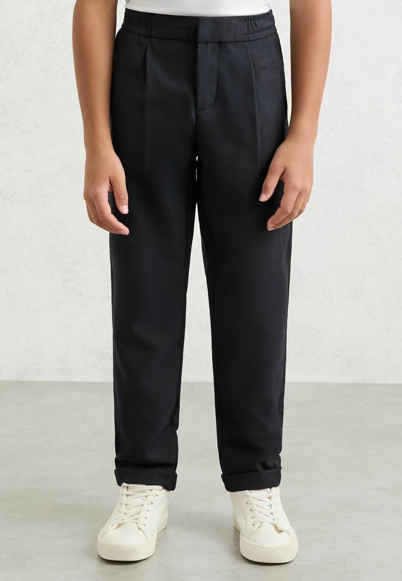 Enfant portant un pantalon noir avec une taille élastique et des baskets blanches, debout sur un sol gris clair contre un mur gris.