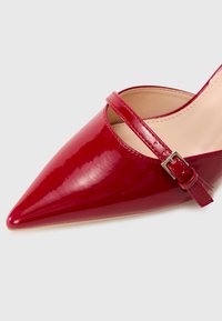 Red patent leather high-heeled shoe with a pointed toe and a single strap secured by a silver buckle. Smooth texture with a glossy finish.