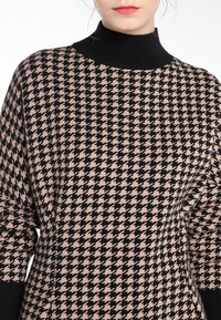 Pull en tricot avec un motif pied-de-poule noir et beige, présentant un col haut en côtes noir, une texture douce et une coupe décontractée.