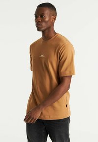 T-shirt marrone a maniche corte con finitura testurizzata, caratterizzato da un piccolo logo bianco sul petto e un sottile dettaglio con spacco laterale.