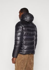 Homme portant une veste en doudoune matelassée noire et brillante avec capuche, de profil, associé à un pantalon en velours côtelé marron clair sur un fond uni.