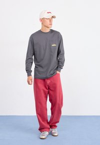 Young man standing indoors wearing a gray long-sleeve shirt, red loose pants, silver sneakers, and a white cap looking to the side.