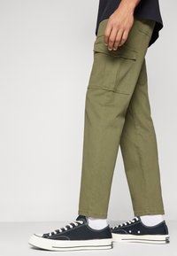 Olivové cargo kalhoty se bočními kapsami, zúženým střihem a hladkým povrchem, kombinované s černými plátěnými teniskami a bílými ponožkami.