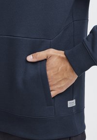 Hanorac bleu marin cu o textură specială, dotat cu o buzunar frontal, manșete ribbed și un mic etichetă de marcă la tiv.