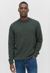 Groen gebreid sweater met een geribbelde ronde hals en manchetten, met meerkleurige spikkels. Gepaarde met blauwe denim jeans.