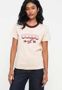 Ροζ βαμβακερό μπλουζάκι με κοντά μανίκια, μπορντό ribbed λαιμόκοψη, και κόκκινο γραφικό κείμενο "GUESS est. 81" μπροστά.