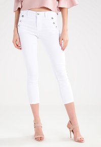 Jean capri blanc ajusté avec cinq poches et des détails de boutons argentés sur le côté. Le matériau semble extensible et lisse.