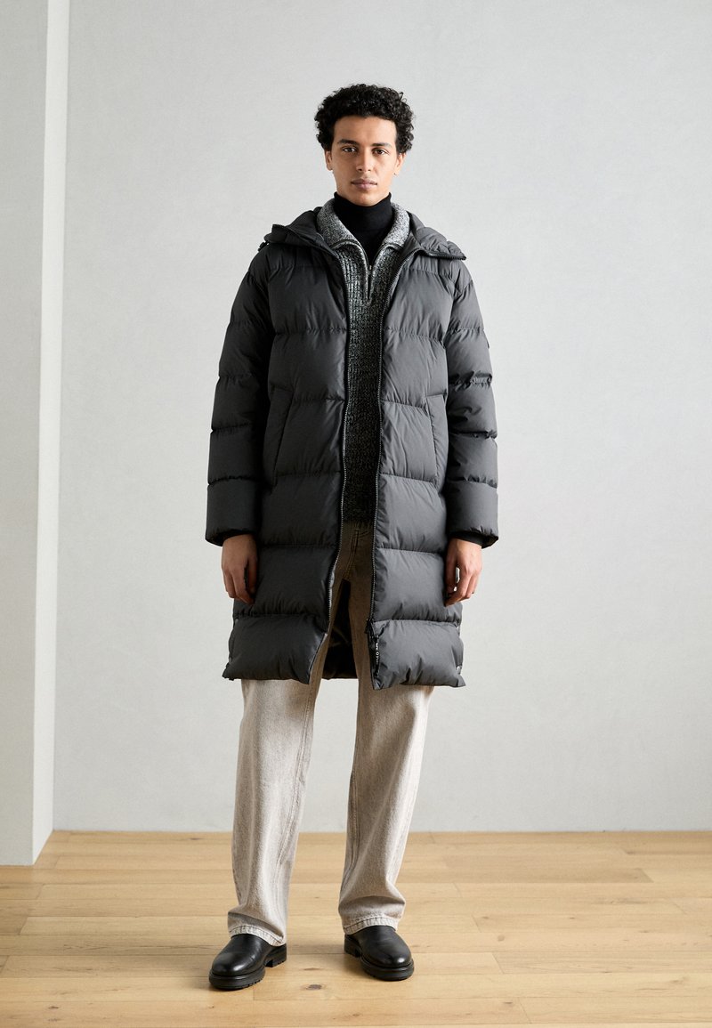 Abrigo puffer negro con diseño acolchado, capucha y cierre de cremallera, usado sobre un suéter oscuro, combinado con pantalones claros y zapatos negros.