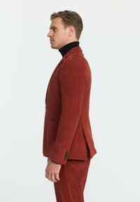 Roter Cordanzug mit einer taillierten Design, vertikalen Rippungen, einem Revers und einem schwarzen Rollkragenpullover darunter.