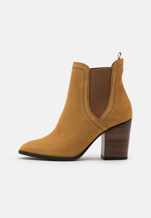 Bottine en daim beige avec panneaux latéraux élastiques, talon bloc en bois, embout pointu et languette à l'arrière.