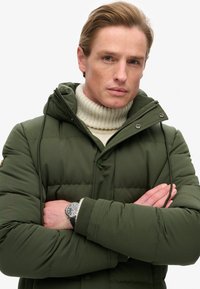 Zielona puchowa kurtka z kapturem na zamek, ściśniętymi mankietami i puszystym białym golfem pod spód. Posiada czarne akcesoria i pikowaną fakturę.