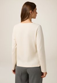 Blusa de manga larga en color crema, fabricada en un tejido suave, con escote cuadrado y un corte holgado. La parte posterior tiene un diseño simple y limpio.