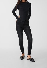 Top negro de manga larga con dobladillo asimétrico, combinado con jeans ajustados oscuros y zapatos planos de punta con adornos decorativos.