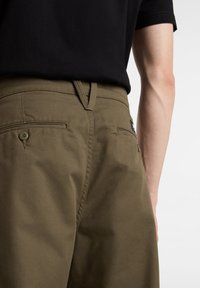 Närbild av en person som bär olivgröna byxor och en svart skjorta, som visar baksidan av fickan och bältesöglorna med detaljerad sömnad.