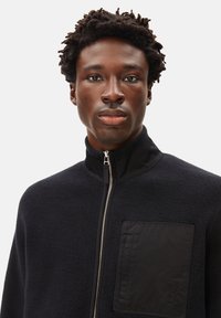 Schwarze Fleecejacke mit hohem Kragen, Reißverschluss und einer quadratischen Brusttasche mit strukturiertem Akzent. Glattes Material mit subtiler glänzender Optik.