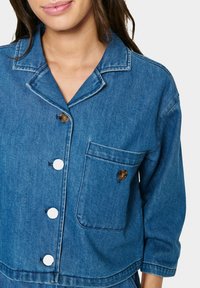 Jean jakna z ovratnikom, rjavimi gumbi, belimi zaponkami, enim žepom na prsih in sproščenim krojem v svetlo modri barvi.