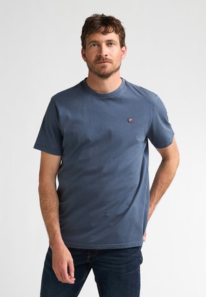 Man met kort bruin haar en baard, die een effen blauw T-shirt en donkere spijkerbroek draagt, staand tegen een witte achtergrond.