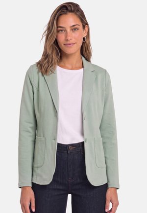 Jonge vrouw met schouderlang haar draagt een lichtgroene blazer over een wit overhemd en donkere, hoog getailleerde spijkerbroek tegen een effen achtergrond.