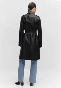 Czarny płaszcz prochowiec z imitacji skóry ze spinką w pasie, pagonami na ramionach i długimi rękawami, zestawiony z jasnoniebieskimi dżinsami o szerokich nogawkach.