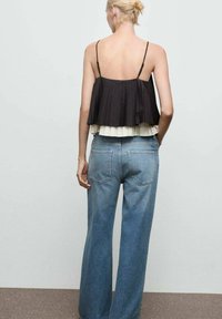 Donna con capelli biondi raccolti in un chignon indossa un top senza maniche plissettato bianco e nero e jeans blu a vita alta, in piedi su uno sfondo neutro.