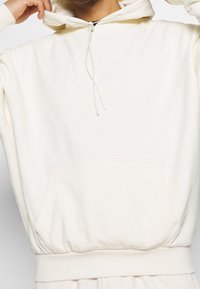 Sudadera color crema hecha de una tela suave, con un bolsillo frontal, capucha con cordón y un logotipo bordado en blanco en el pecho.