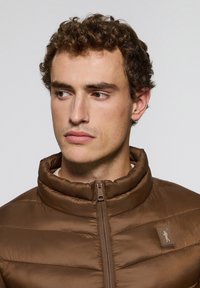 Chaqueta de plumas marrón con un cuello alto, costuras horizontales y un cierre de cremallera. Presenta un pequeño logo en el pecho y una textura lisa y brillante.