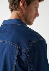 Chaqueta de mezclilla en azul oscuro, con un cuello clásico, costuras en contraste y una pequeña etiqueta cuadrada en la parte posterior. Tejido texturizado.