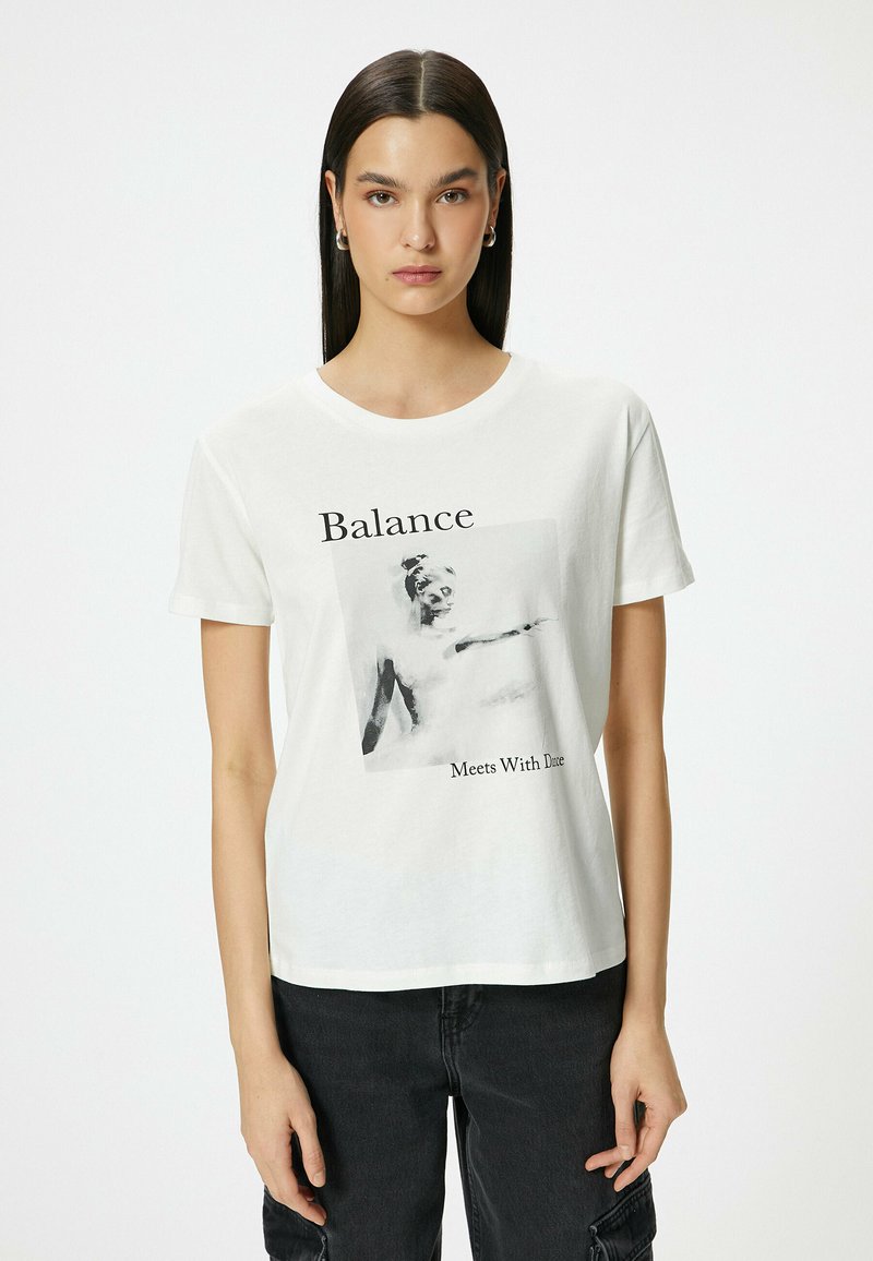Hvid bomulds T-shirt med fotoprint af en figur med teksten "Balance Møder Døden" i sort. Korte ærmer og rund hals.