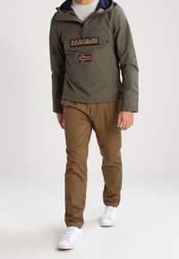 Uomo che indossa una giacca Napapijri verde con una toppa della bandiera norvegese, pantaloni marroni e scarpe da ginnastica bianche mentre cammina su uno sfondo neutro.
