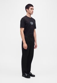 Jeune homme debout de profil, portant un t-shirt noir avec un logo, un pantalon noir à taille élastique et des chaussures habillées noires sur fond blanc.