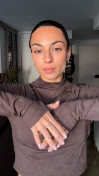 Langærmet, figursyet brun top med høj krave, blød tekstur og tommelfingerhuller ved ærmerne, kombineret med negle i neutrale toner og synlige tatoveringer.