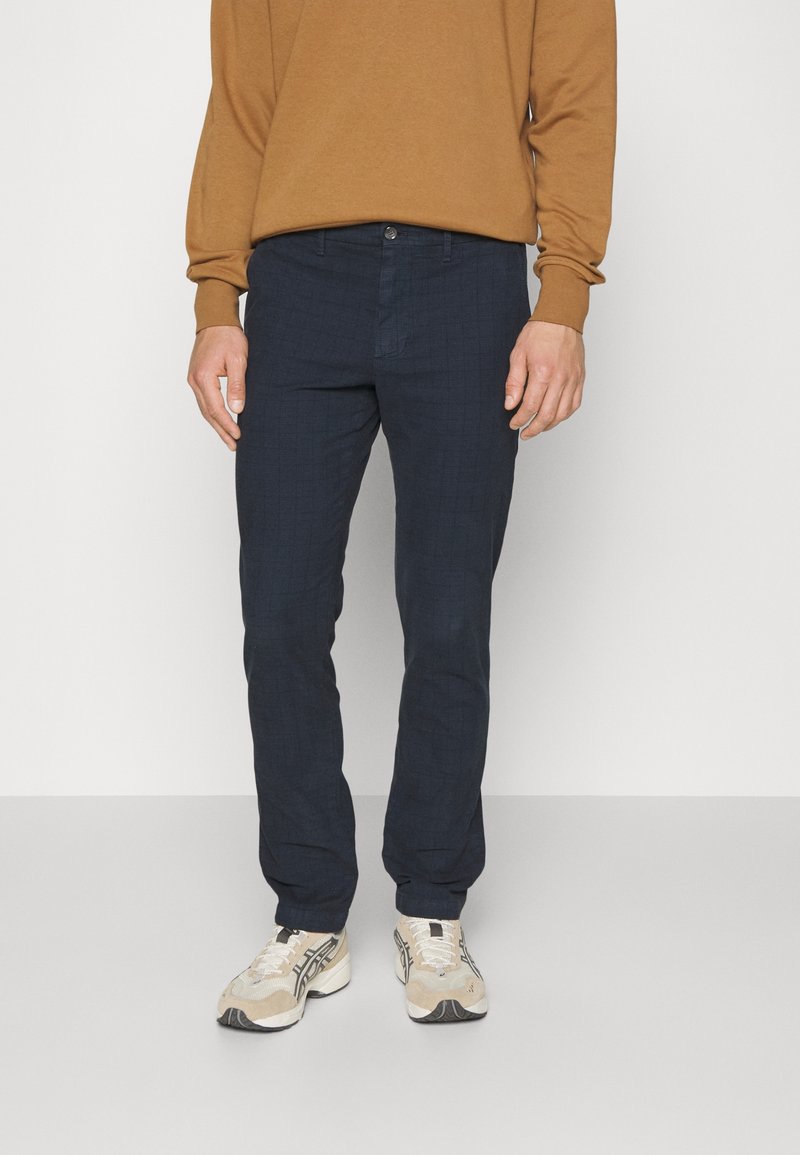 Marinblå rutiga byxor i vävt tyg, med rak benform, framfickor och knäppning med knapp, tillsammans med beige sneakers.