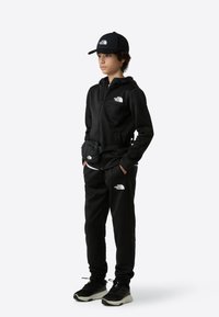 Čierna bunda na zips a nohavice z mäkkej látky, doplnené čiernou čiapkou a malou ľadvinkou. Výrazné biele logo akcenty.